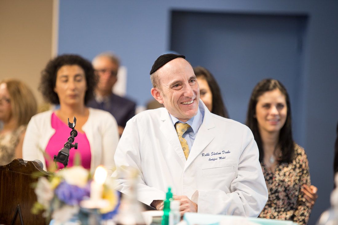 Rabbi Shalom Denbo, in a white coat embroidered with his name, smiling at a ceremony